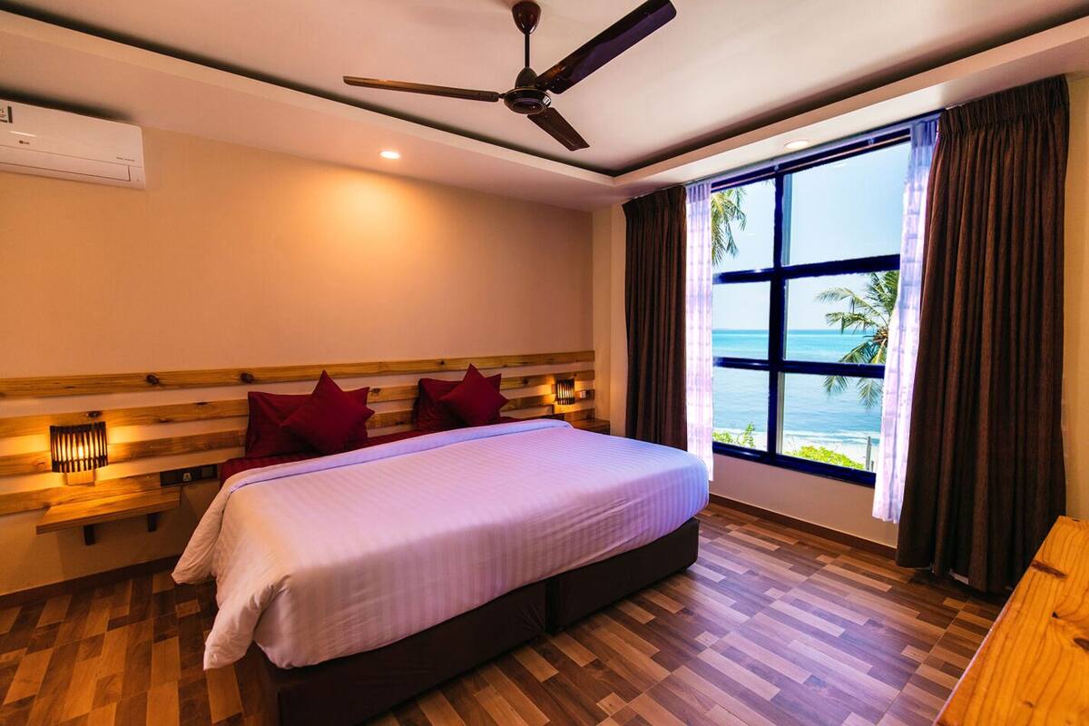 A hotel room with a large bed, wooden headboard, a ceiling fan, a window with a view of the sea, and a wooden floor.