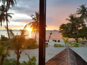 Vista da praia com palmeiras, uma estrutura de telhado de palha e um pôr do sol sobre o oceano.