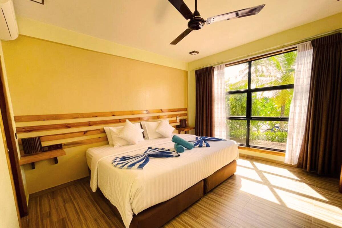 A hotel room with a wooden headboard, a bed with white linens, a ceiling fan, and a large window with curtains.