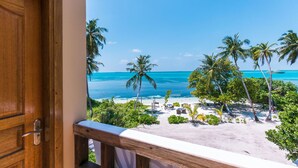 Una porta in legno che dà su un balcone con vista su una spiaggia, palme e un mare blu e limpido.