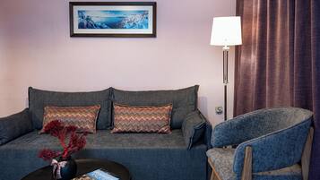A living room with a grey sofa, patterned cushions, a blue armchair, a black coffee table with books, a vase with red branches, a framed picture on the wall, and a floor lamp.