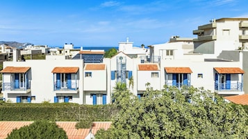 A row of white buildings with blue shutters and orange roofs, likely residential apartments, with a clear view of the sea in the background.