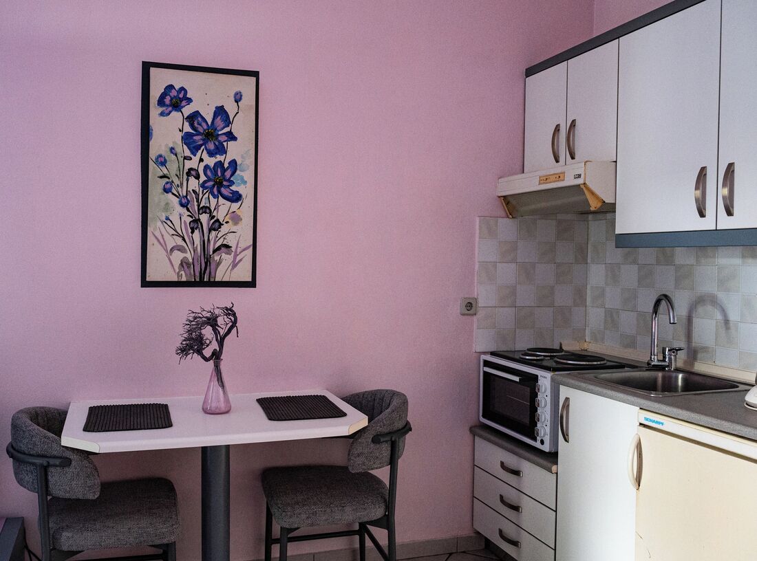 A compact kitchen with a white countertop, a microwave, and a sink. There is a framed floral artwork on the wall and a vase with a plant on the table.