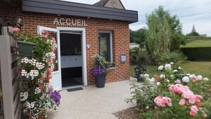 A brick building with a sign reading "ACCUEL" and a door open. There are flower pots with various flowers outside. - Le Camp des Roses (Aubers)