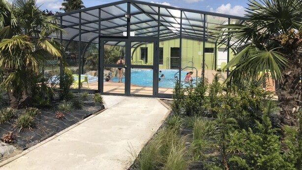 A covered pool area with a glass enclosure, surrounded by tropical plants and a concrete pathway. - Le Camp des Roses (Aubers)