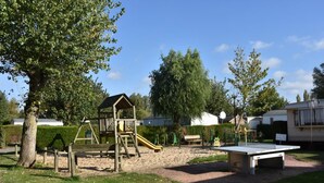 A playground with a slide, climbing structure, and a table tennis table in a park area with trees and a clear sky. - Le Camp des Roses (Aubers)