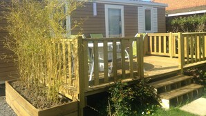 A wooden deck with a railing, a small garden area with bamboo, and a house with a brown siding exterior. - Le Camp des Roses (Aubers)