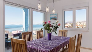 Zona de menjador amb taula preparada per a quatre, mantell morat i un ram de flors. Hi ha finestres grans que ofereixen vista al mar i a edificis.