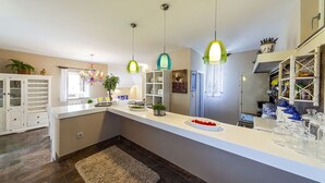 A modern kitchen with white cabinetry, a large island, hanging pendant lights, and a variety of kitchenware. - Villa Skalinada (Baska Voda)