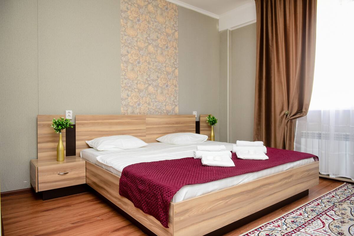 A neatly made bed with a red blanket, wooden headboard, and two bedside tables with vases.