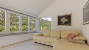 A spacious room with a beige sofa, large windows offering a view of greenery and hills, and a framed painting on the wall. - Villa Roglic (Zagvozd)