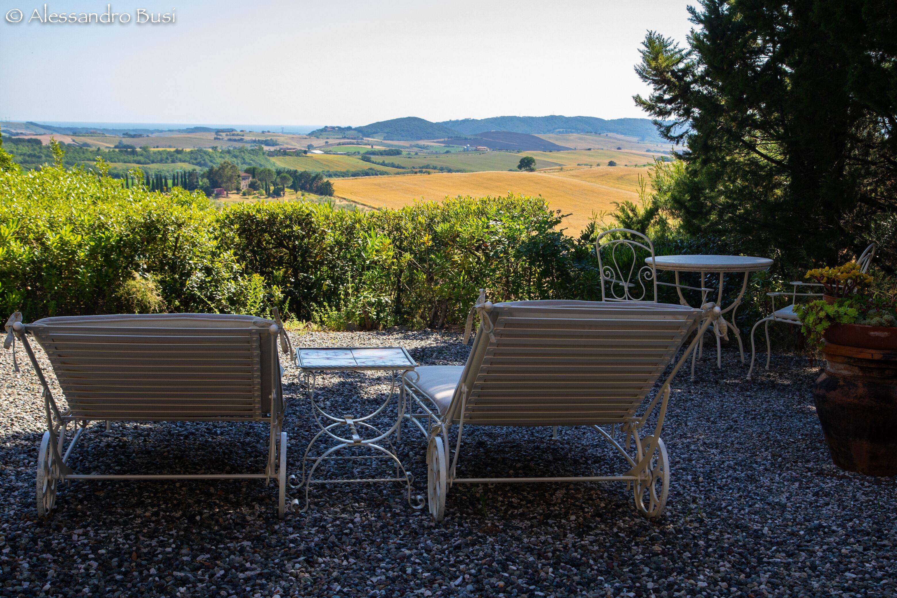 Un patio con dos sillas de metal y una pequeña mesa, rodeado de grava y follaje, con vistas a un paisaje pintoresco.