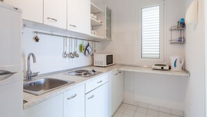 A compact kitchen with white cabinets, a stainless steel sink, a microwave, and a window with blinds. - Holiday home Duje (Okrug)