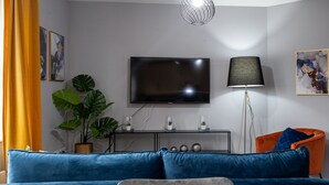 A modern living room with a large flat-screen TV, a floor lamp, a blue sofa, and a small orange chair. - Trafford Place (Manchester)