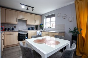 A modern kitchen with light wood cabinets, a white countertop, and a dining table with grey chairs. - Trafford Place (Manchester)