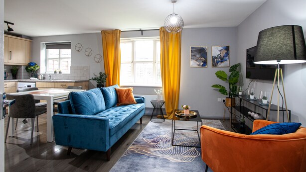 A modern living room with a blue sofa, an orange armchair, and a dining area with a white table and chairs. - Trafford Place (Manchester)