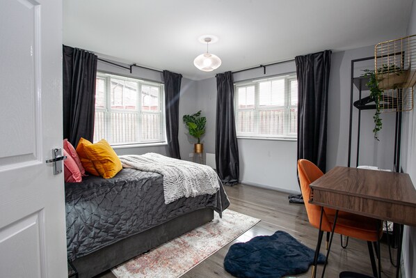 A bedroom with a bed, a desk, and a chair. There are windows with curtains, a hanging plant, and a light fixture. - Trafford Place (Manchester)
