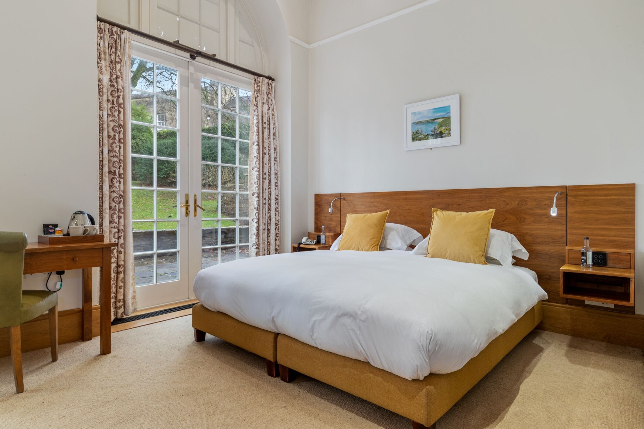 Ein Schlafzimmer mit einem großen Bett, einem Holzkopfende, Nachttischen, einem Schreibtisch und Blick durch eine Glastür.