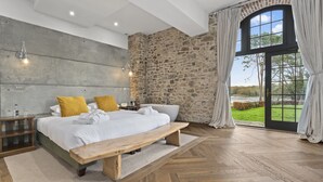 Une chambre avec un grand lit, un banc en bois, un mur en pierre, une baignoire et une vue sur l’extérieur.