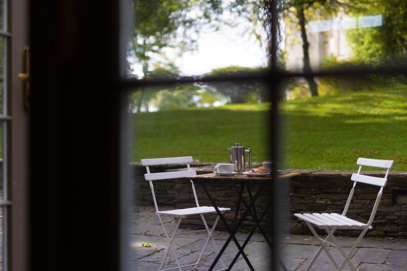 A view through a window of a patio area with a table set for two, a coffee pot, and a plate with a pastry.