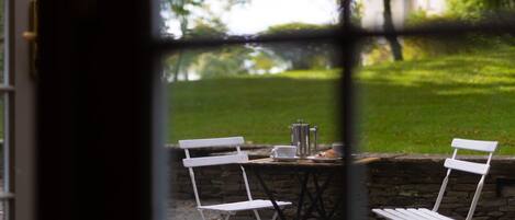 Vista a través de una ventana de un patio con una mesa dispuesta para dos, una cafetera y un plato con un pastel.