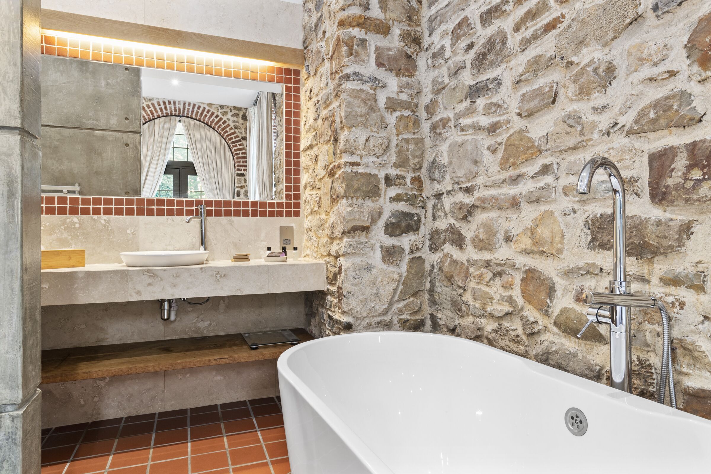 Un baño con pared de piedra, una bañera blanca y un espejo sobre el lavamanos.