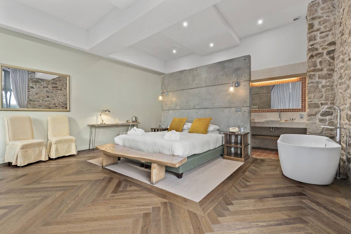 Een moderne slaapkamer met een groot bed, twee stoelen, een bankje, een accentmuur van steen, een grote spiegel en een ligbad.
