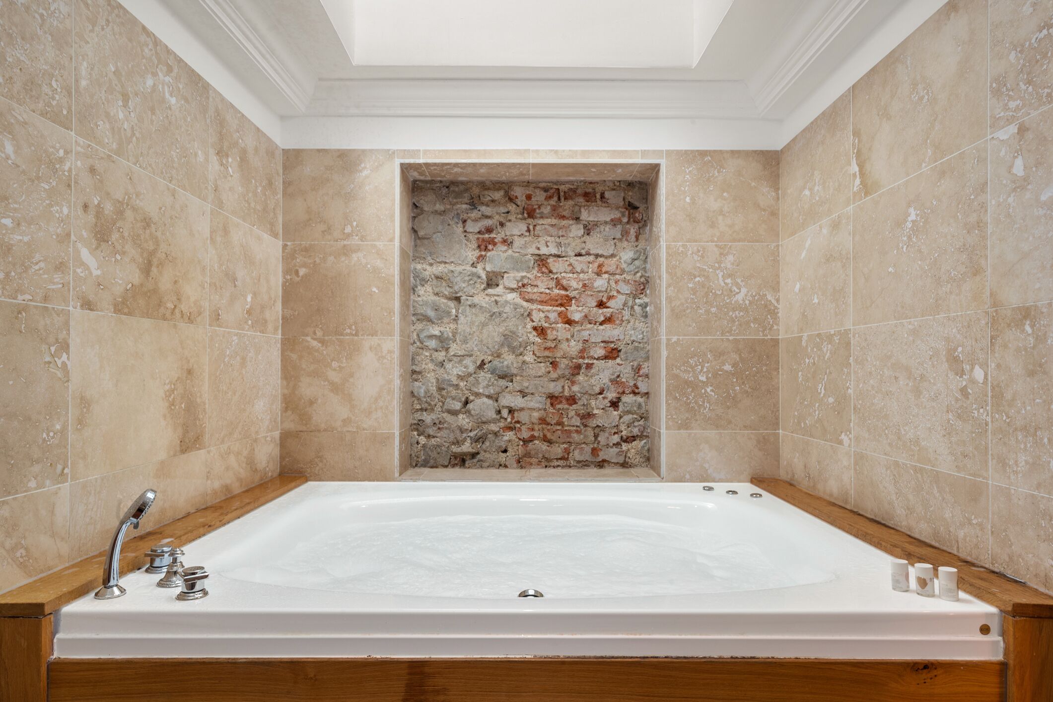 Eine große, rechteckige Badewanne mit Holzrand, vor beige gefliesten Wänden mit einer Ziegelwand in der Mitte.