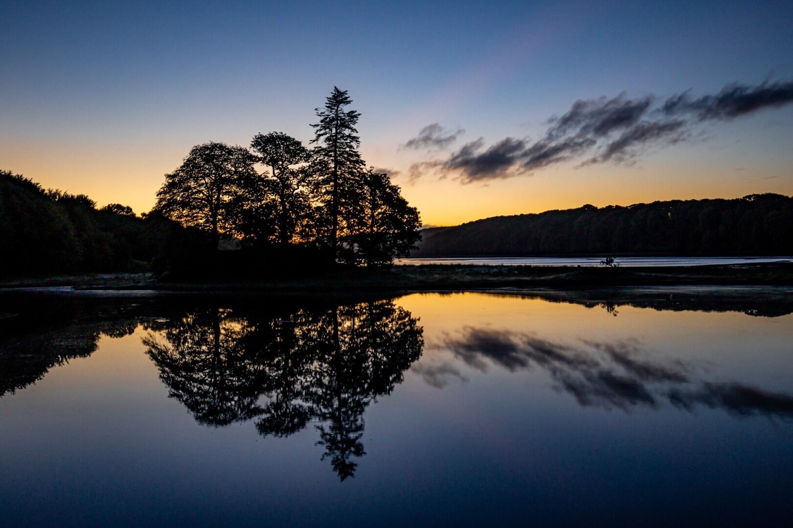 A serene lakeside scene with a calm body of water reflecting trees and a sunset sky.