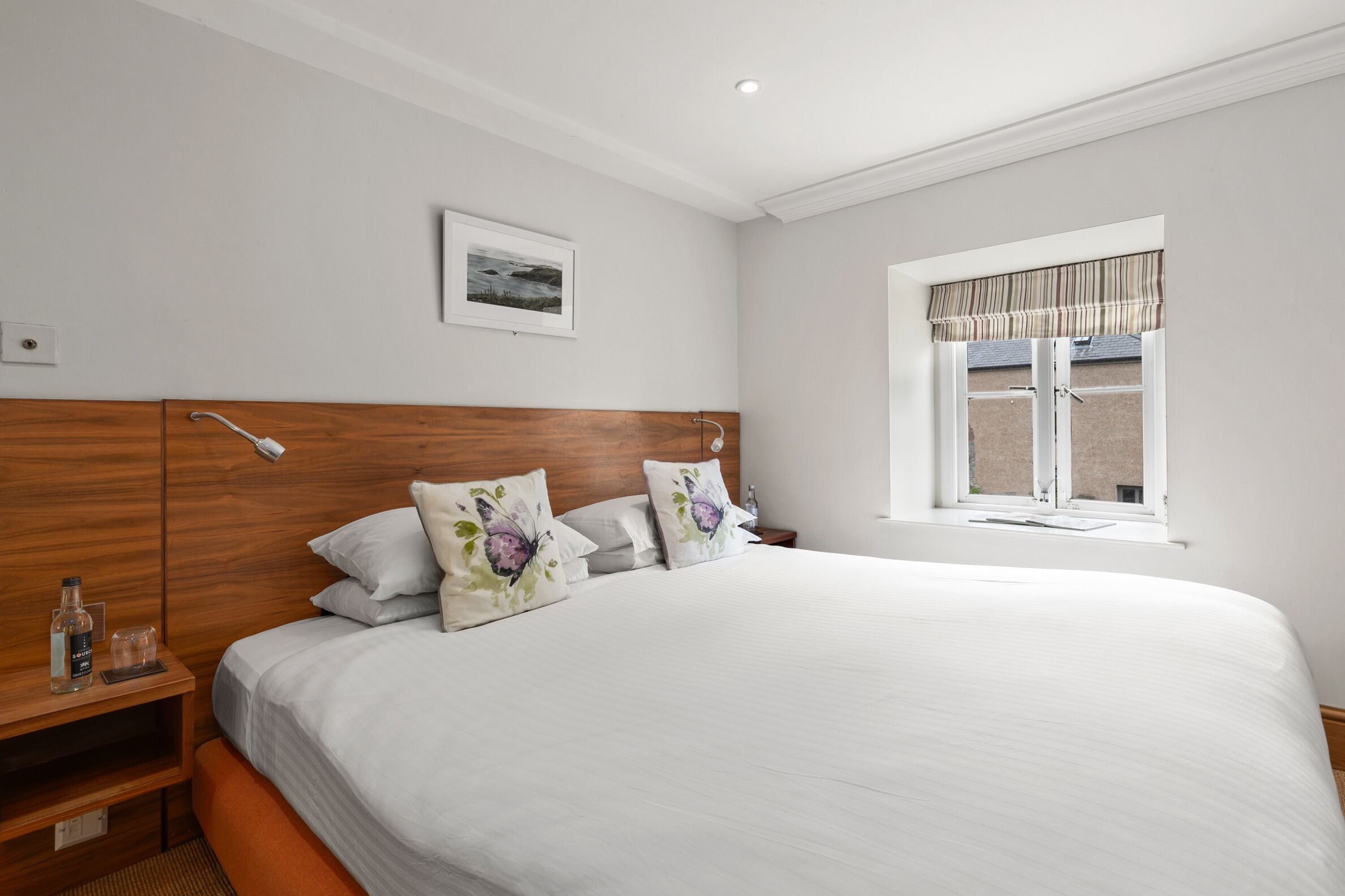 A neatly made bed with floral pillows, a wooden headboard, a bedside table with a bottle and glass, and a window with a striped valance.