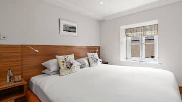 Una cama bien hecha con almohadones florales, cabecera de madera, mesita de noche con una botella y un vaso, y una ventana con cortina a rayas.