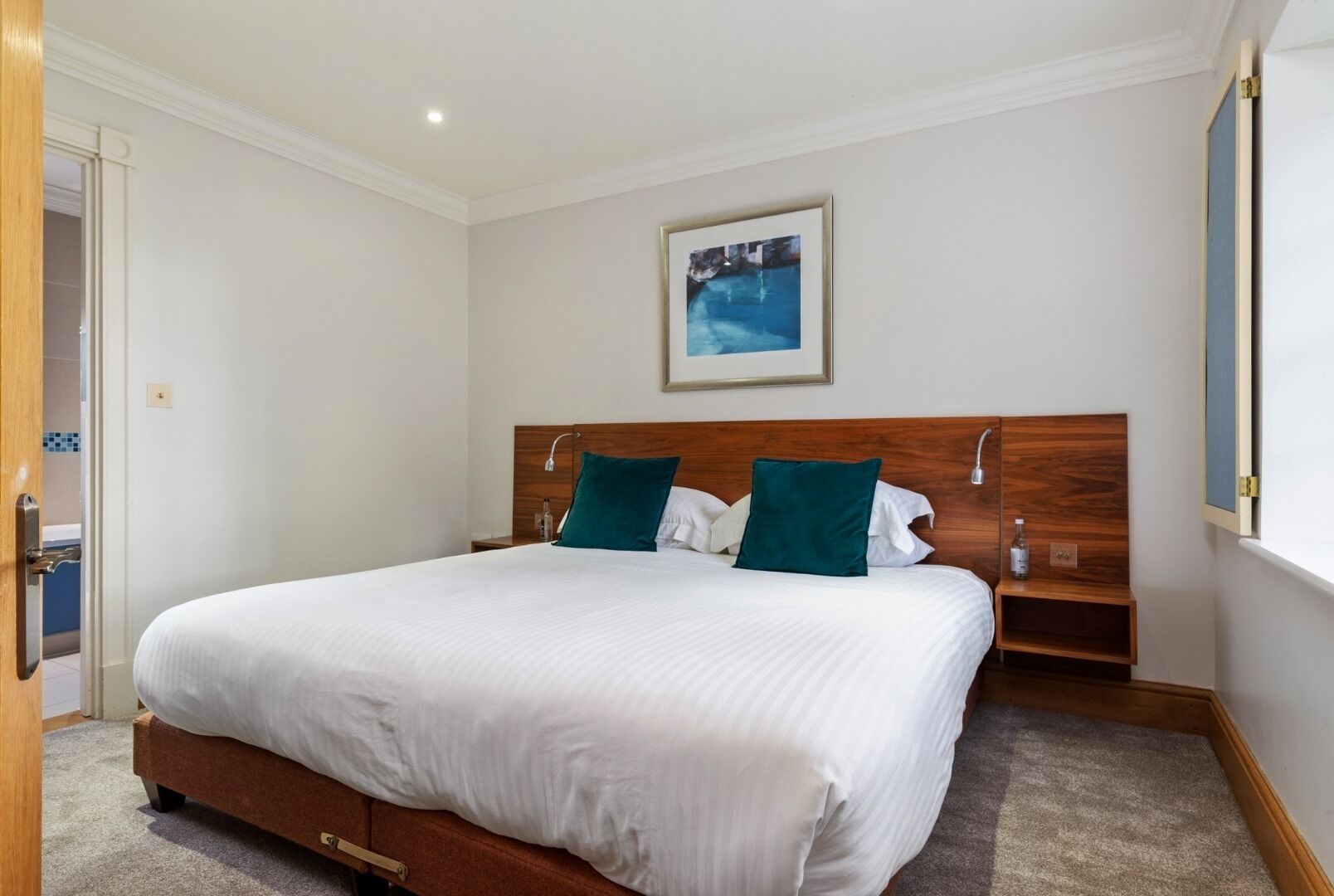 Habitación de hotel con una cama grande, cabecera de madera, dos almohadas, una mesita de noche, un cuadro en la pared y una puerta a la izquierda.