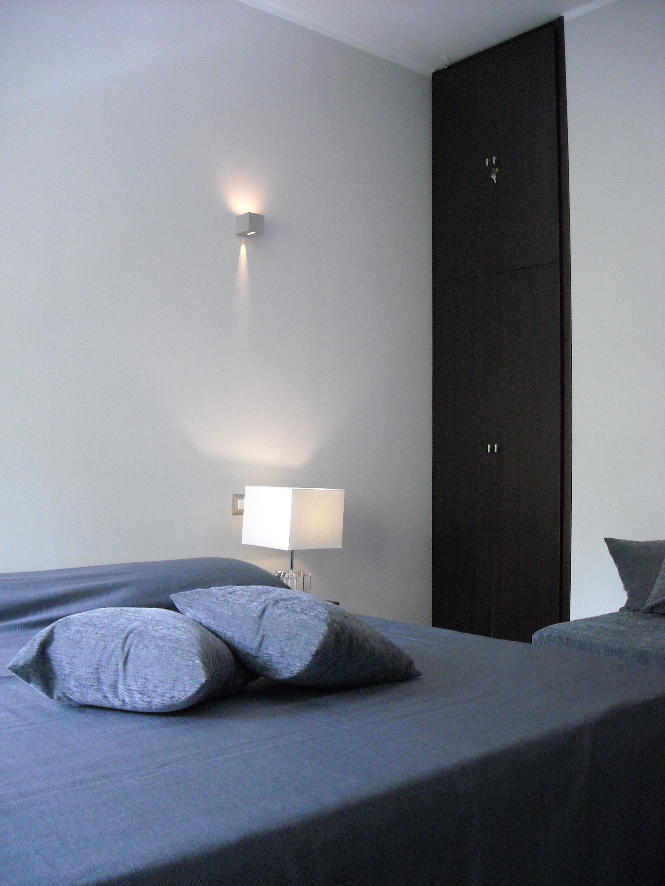 Un dormitorio con cama, almohadas, mesita de noche, lámpara y armario.