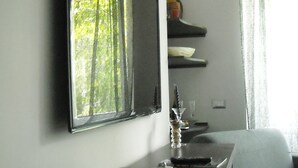 Habitación moderna con televisión de pantalla plana en la pared, una mesita con una bandeja de pasteles, una botella de agua y un vaso. Se observa un equipo de aire acondicionado en la pared y una ventana con cortinas.