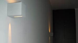 Una habitación con pared blanca, puerta oscura y una pequeña luminaria en la pared.