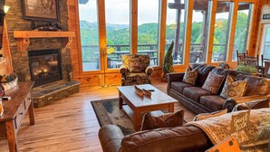 A cozy cabin interior with a stone fireplace, leather furniture, and a large window overlooking a forested mountain.