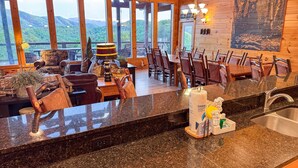 A wooden cabin interior with a dining area, kitchen counter, and large windows overlooking a forested landscape.