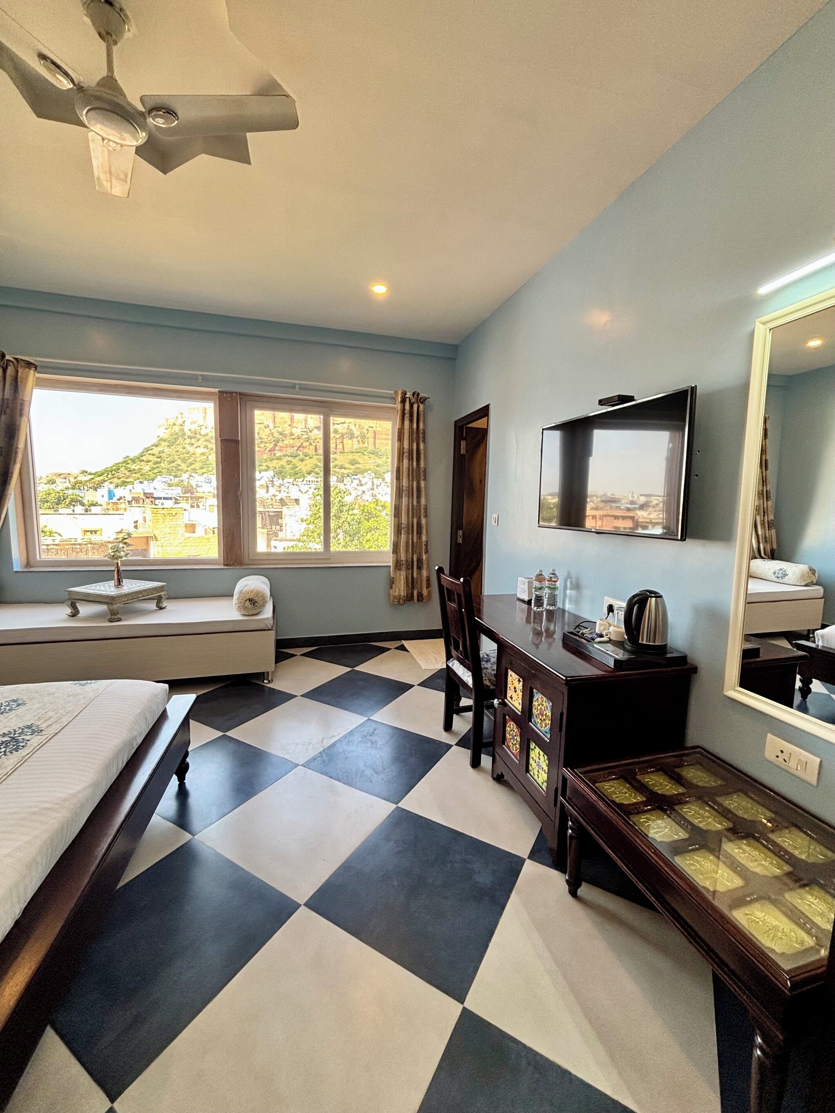 Quarto de hotel com cama, escrivaninha, cadeira, televisão, espelho, janela com vista para uma colina e chão quadriculado.