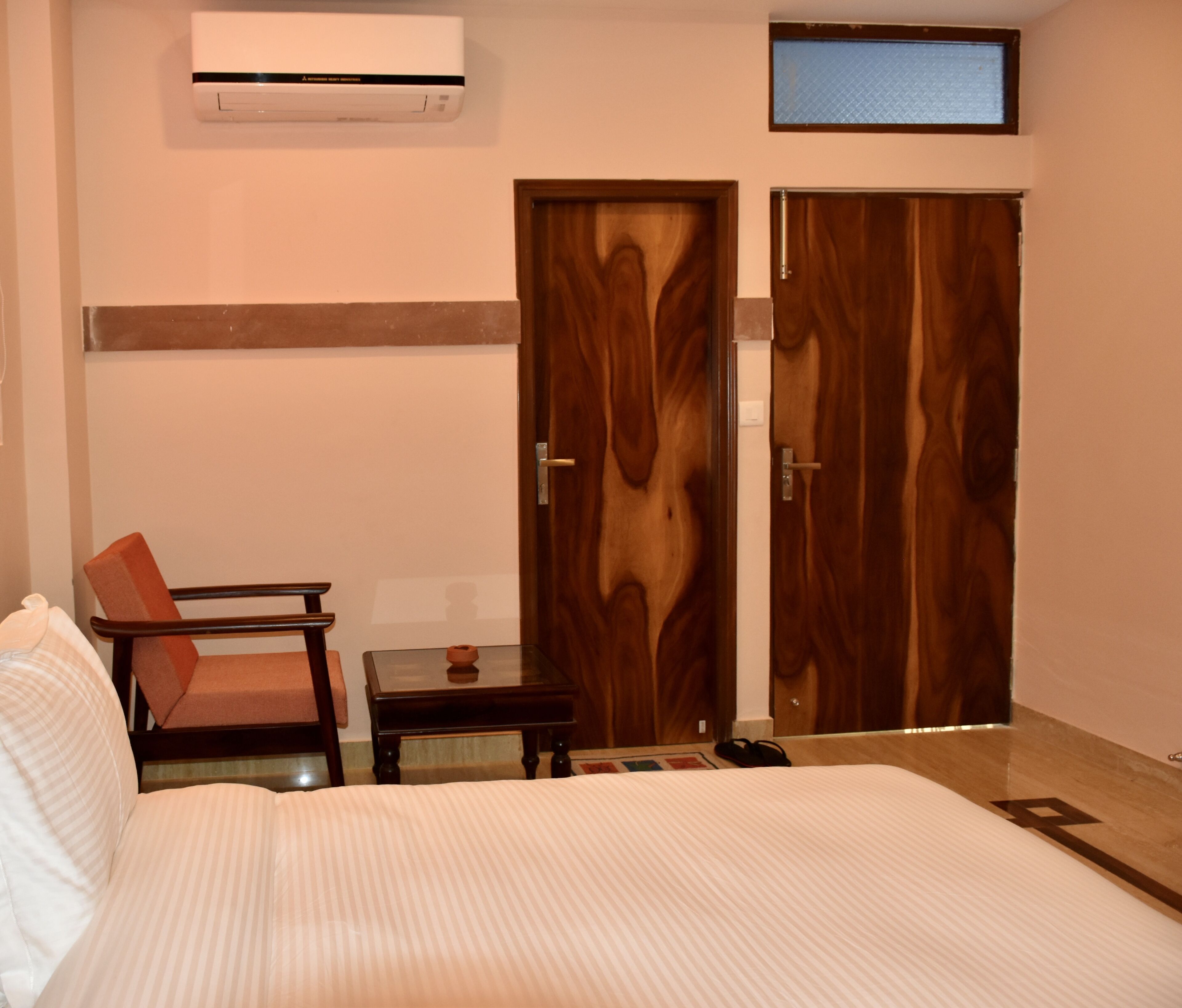 A hotel room with a bed, a chair, a small table, a TV mounted on the wall, and two wooden doors.
