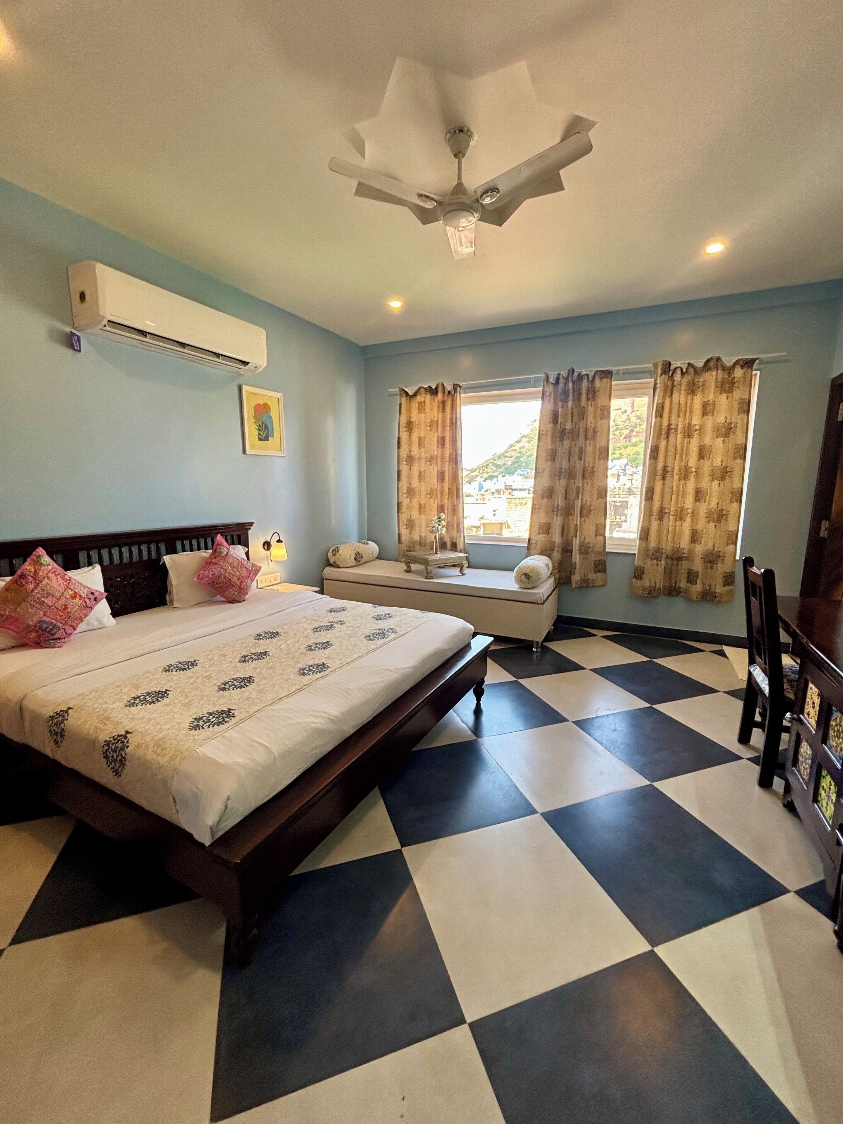 A bedroom with a bed, a bedside table, a chair, a window with curtains, a ceiling fan, and a checkered floor.