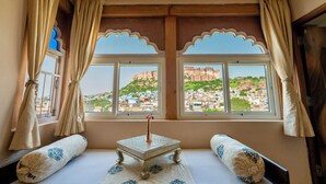 Ein Zimmer mit einem Bett, einem kleinen Tisch mit einer Vase und Blick auf eine Stadtlandschaft durch zwei mit Vorhängen versehene Fenster.