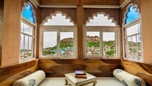 Una habitación con vistas a una colina y edificios, una pequeña mesa con libros, y dos almohadones decorativos.