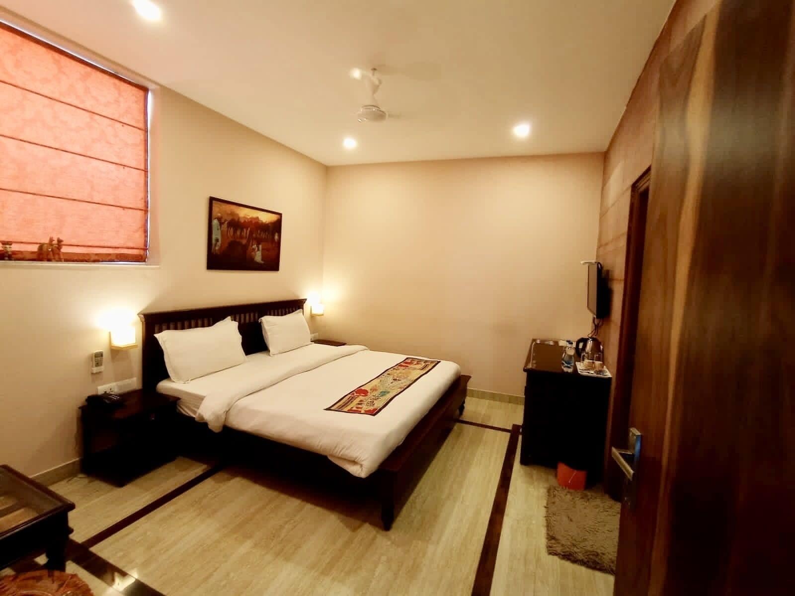 Un dormitorio con cama, mesitas de noche, un televisor montado en la pared y una ventana con persianas.