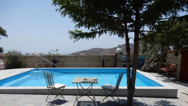 A rectangular swimming pool with a clear blue water, surrounded by stone tiles. There are two metal chairs and a small table with a cushion. A tree is in the foreground, and a stone wall with a gate is in the background. - Palmera, quiet studio with terrace and view to the mountains (Las Zocas)