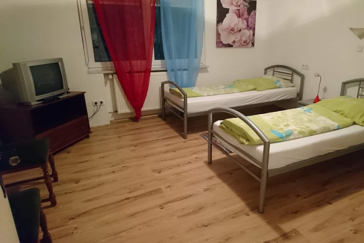 Une petite pièce avec deux lits superposés en métal, un plancher en bois, un téléviseur posé sur un meuble en bois et une fenêtre ornée de rideaux légers.