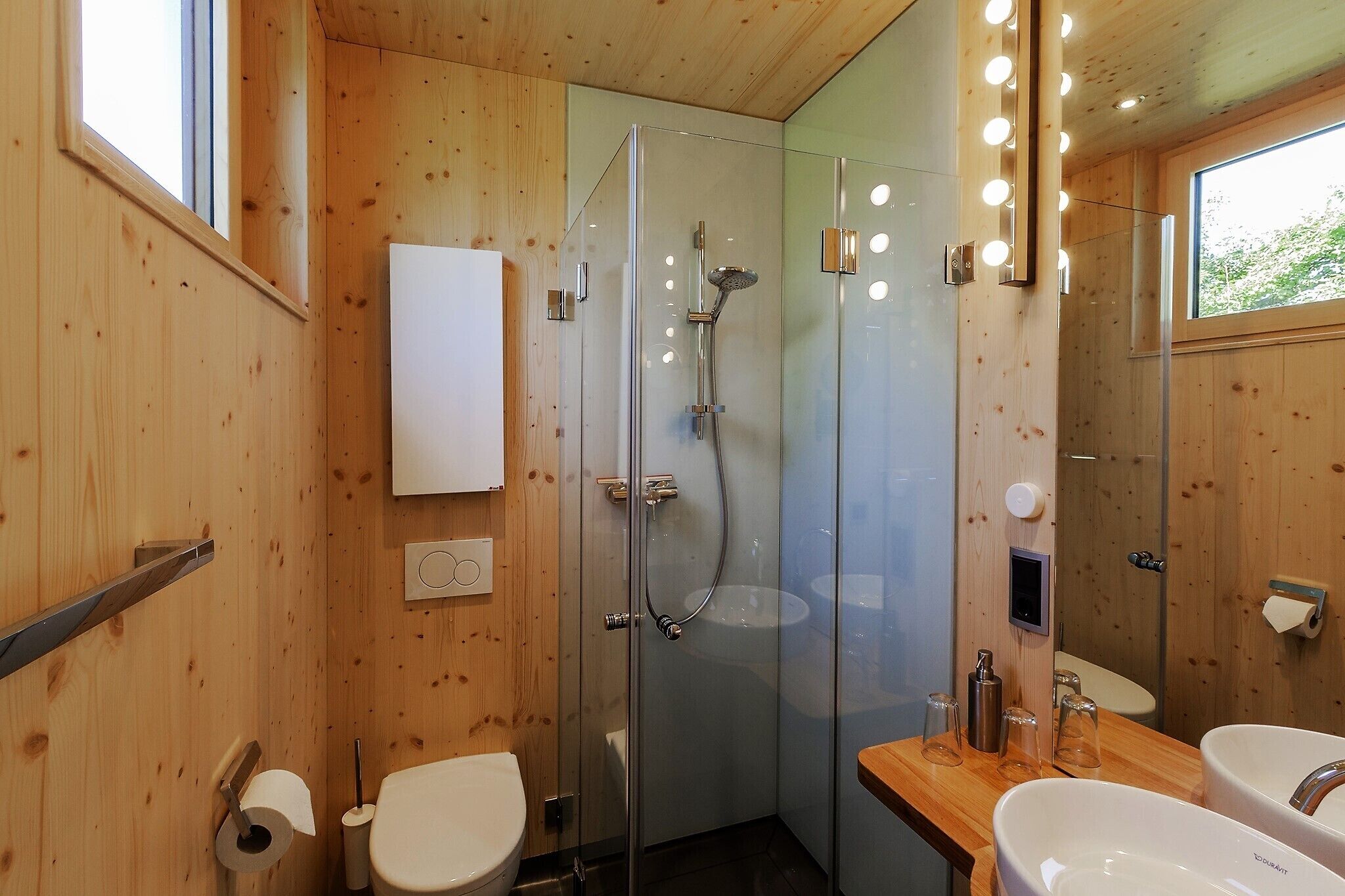 A wooden-paneled bathroom with a glass shower enclosure, two sinks, and a mirror with lights.