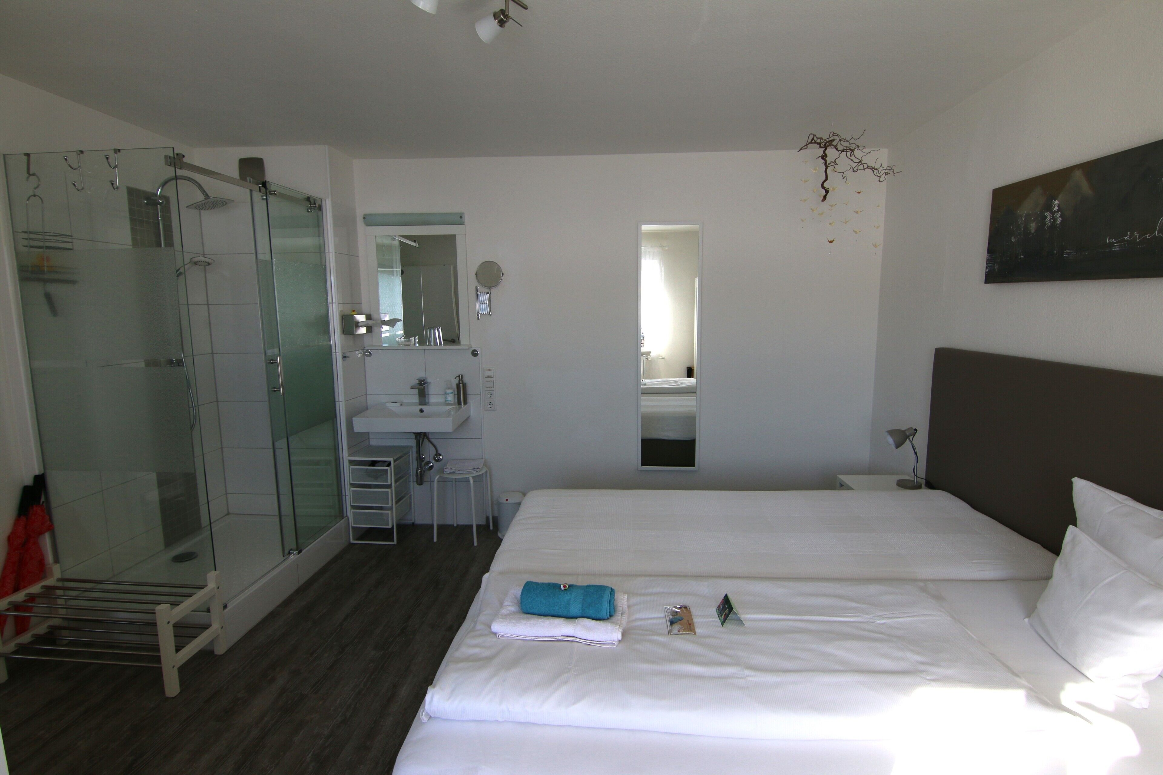 A hotel room with a glass-enclosed shower, a bed with white linens, a small desk, and a nightstand.