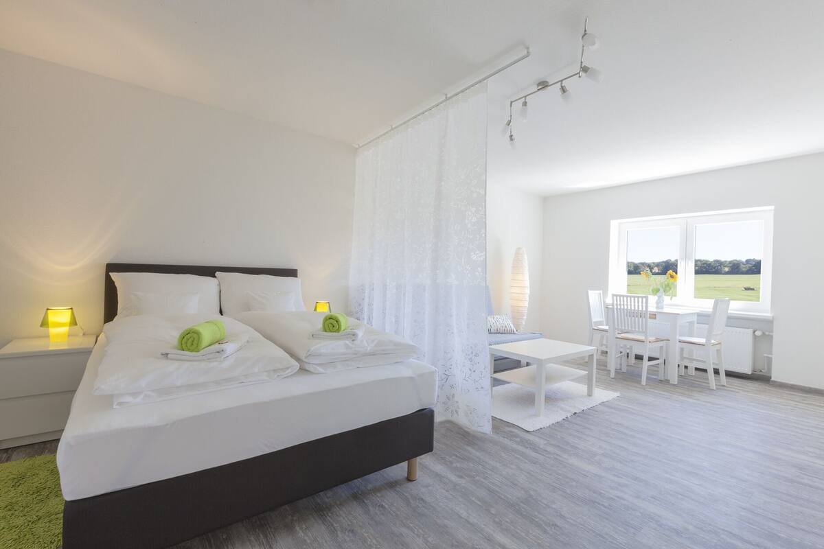 Quarto com cama, duas mesas de cabeceira com abajures, escrivaninha com cadeiras e janela com vista para o exterior.