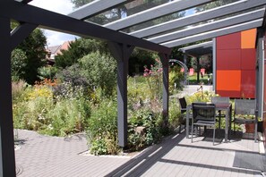 Un espacio al aire libre con patio, una pérgola, una mesa con sillas y un jardín con diversas plantas y flores.
