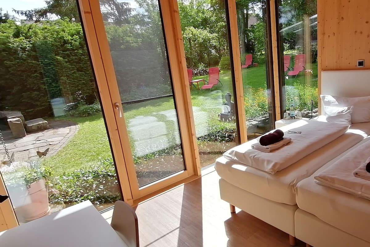 Quarto com uma grande porta de vidro deslizante que dá para um jardim, cama com roupa de cama branca e teto de madeira.